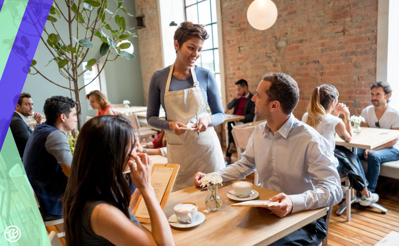 A server in an apron smiling while taking an order from a couple at a table in a busy, sunlit restaurant.