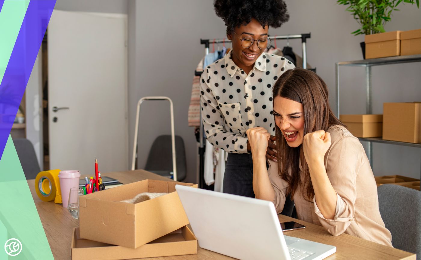 Two colleagues celebrating in a workshop or office setting as they look at a laptop together, with shipping boxes and packing materials in the background.
